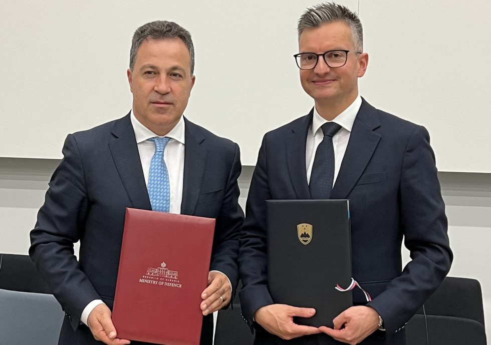 Na levi strani albanski minister za obrambo drži rdečo mapo, na desni strani pa slovenski obrambni minister drži črno mapo z zlatim grbom Slovenije.