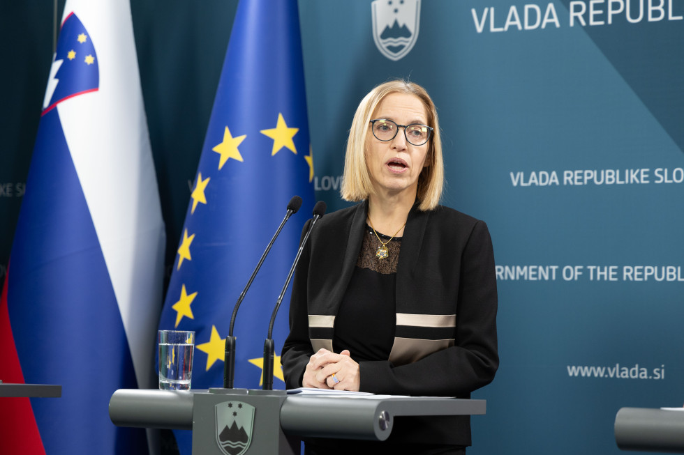 Woman standing behind a lectern during a press conference, surrounded by the flags of Slovenia and the European Union. The background features the logo of the Government of the Republic of Slovenia.