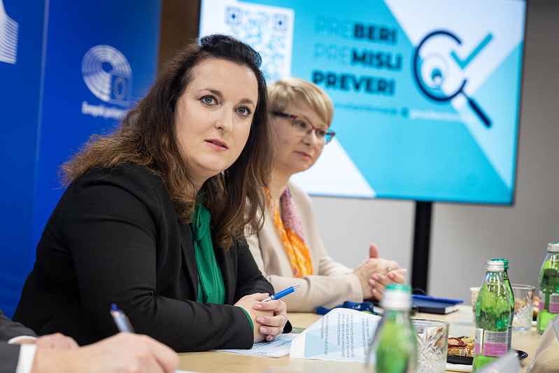 A professional meeting with two women sitting at a table, taking notes. A screen in the background displays a message in Slovenian: "Preberi, Premisli, Preveri." Bottled water and documents are on the table.