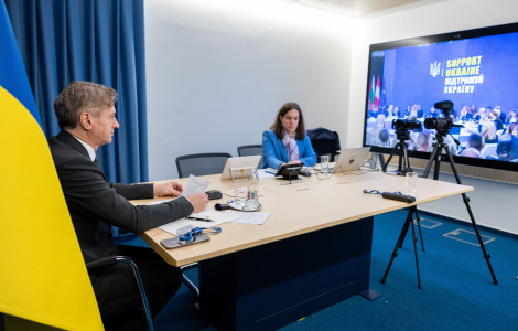 PV Ukrajina 1 24022025 (Robert Golob and his assistant seat at a table, with a flag and a curtain on the left and a screen on the right.)