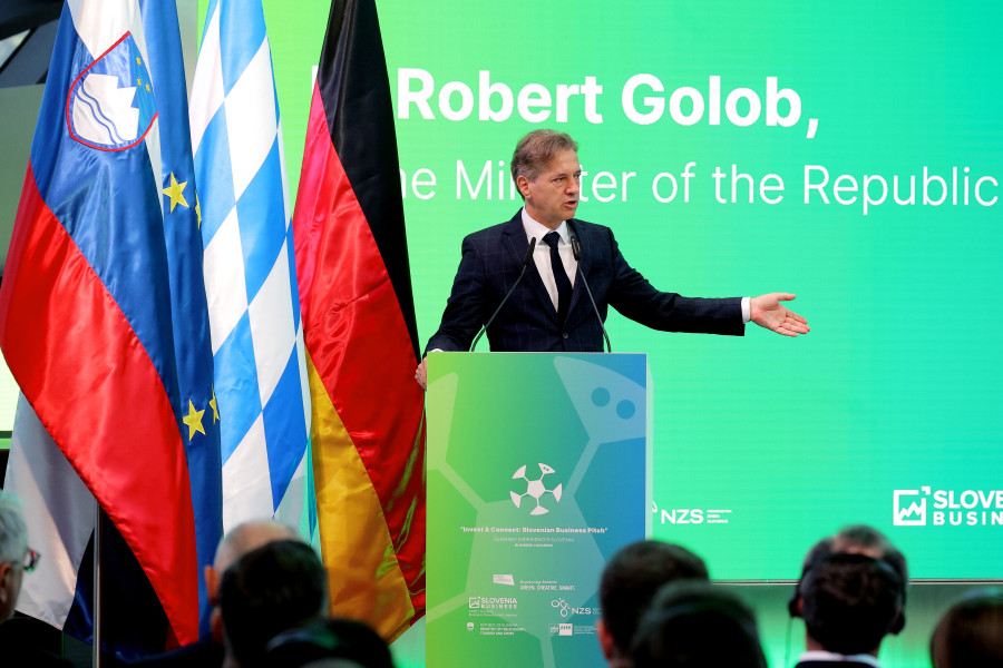 Predsednik vlade med govorom stoji za govorniškim pultom, za njim zelen pano, ob njem slovenska, bavarska in nemška zastava