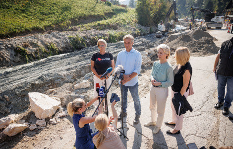 Crna 12 (The Prime Minister, the President of the European Commission and the Mayor of Črna na Koroškem stand in front of the journalists' microphones at the road reconstruction section. Behind them are piles of sand and construction machinery and workers.)