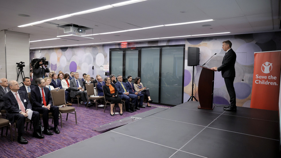 The Prime Minister stands behind the lectern and addresses the participants sitting in the hall. Behind him, on the wall, is a red sign with the words Save the children