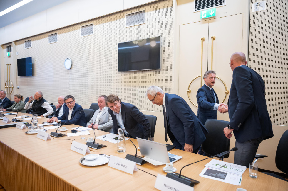 In the meeting hall, the Prime Minister shakes hands with the representatives of the automotive industry, who are sitting at the table. There are name plates on the table.