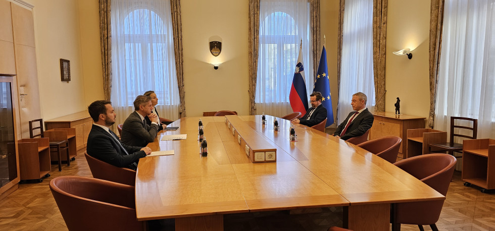 Five people are seated at a large table, engaged in conversation. In the background, the Slovenian and European flags are displayed.