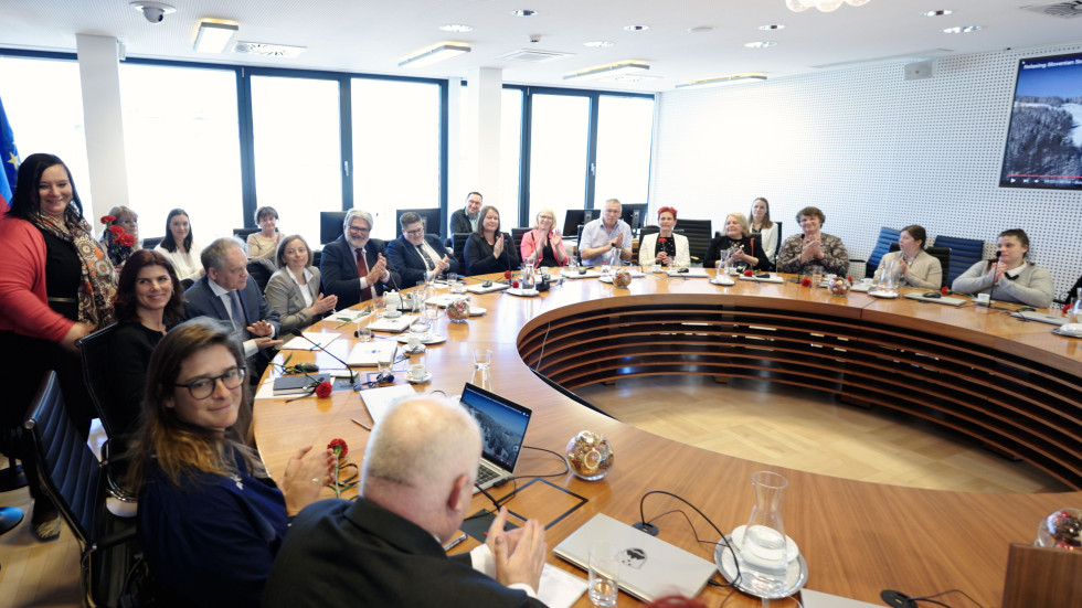 Skupina ljudi sedi okoli okrogle mize v konferenčni sobi, ploskajo in se pogovarjajo. Na mizi so prenosniki, mikrofoni, skodelice kave in rdeče rože.