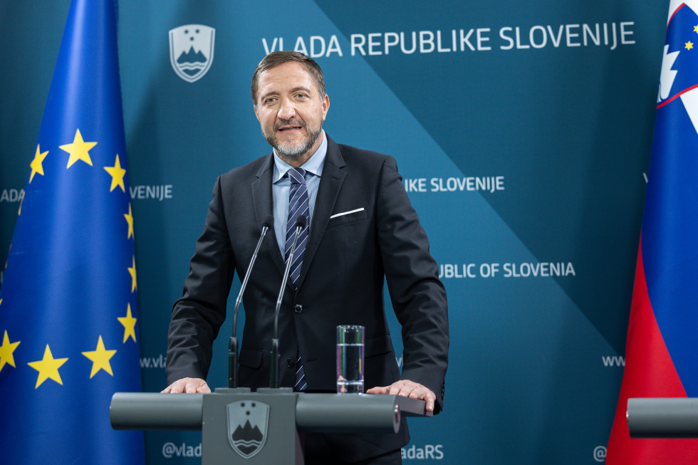 Minister za finance stoji za govorniškim pultom, na katerem je mikrofon in kozarec z vodo. Za njim sta slovenska in evropska zastava in ozadje z napisom Vlada Republike Slovenije in grb Republike Slovenije.