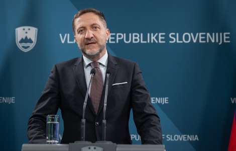 20250130 01410263 (Bradat moški v obleki in kravati govori na tiskovni konferenci. V ozadju je modra stena z napisom "Vlada Republike Slovenije" in slovenskim grbom.)