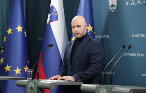 20250305 01419231 (A bald man with a serious expression speaks at a press conference, standing behind a podium. He is dressed in a dark blue blazer and a black turtleneck. Behind him are the Slovenian and European flags, as well as the official inscriptions of the Governmen)