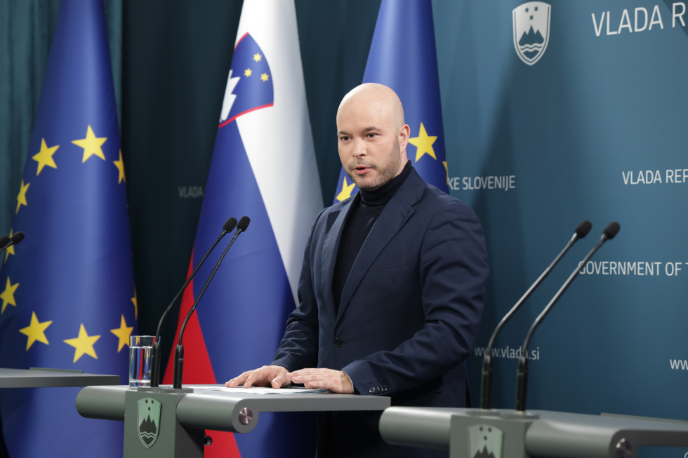 A bald man with a serious expression speaks at a press conference, standing behind a podium. He is dressed in a dark blue blazer and a black turtleneck. Behind him are the Slovenian and European flags, as well as the official inscriptions of the Governmen