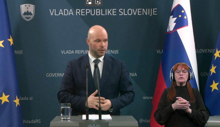 The image shows a press conference with a man speaking at a podium, with the backdrop displaying "Vlada Republike Slovenije" (Government of the Republic of Slovenia). The Slovenian and European Union flags are visible in the background. A sign language in