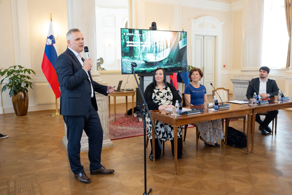 Moški v obleki stoji z mikrofonom v roki, desno od njega za mizo sedita ženski.