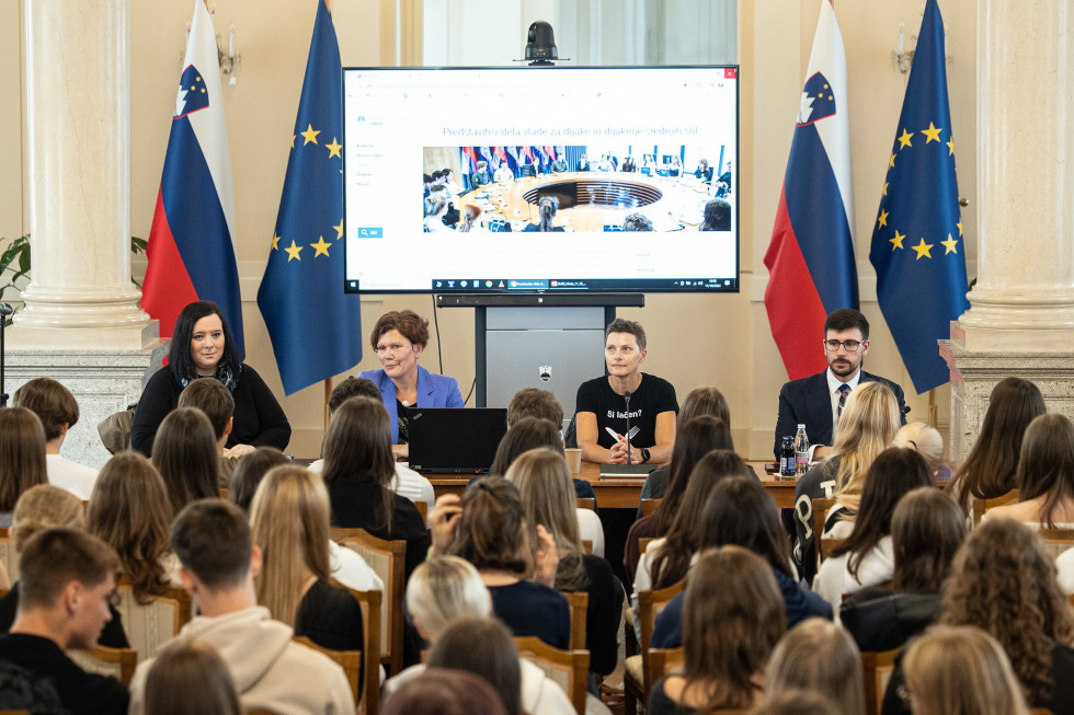 Dijaki sedijo v polni dvorani in gledajo na govorniški pult. Ženska, oblečena v črni majici in hlačah stoji, poleg nje velik ekran.