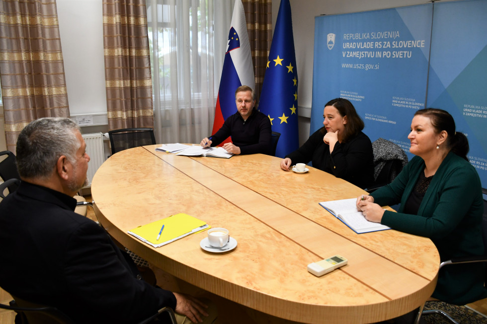 Državna skretarka s sodelavcema z Josephom Valencicem. Sedijo za mizo, v ozadju plakat urada in slovenska in evropska zastava.