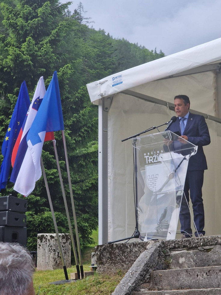 Minister Matej Arčon med govorom za govornico na odru. Na levi strani slovenska, tržiška in evropska zastava. 