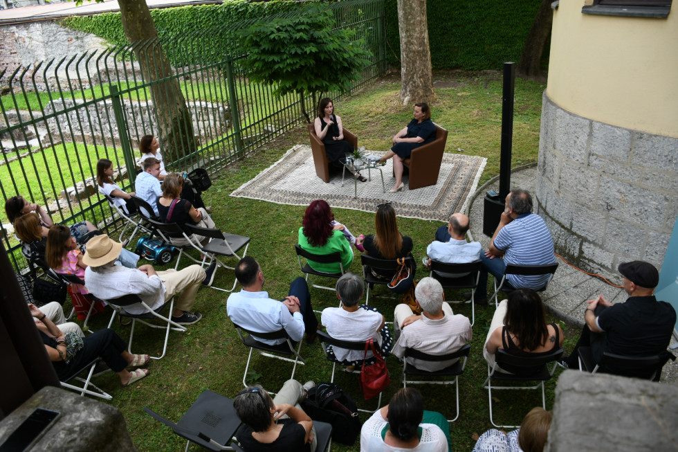 Lucija Stupica in Vesna Humar sedita na preprogi sredi vrta obdanega z drevesi. Pred njima udeleženci prireditve.