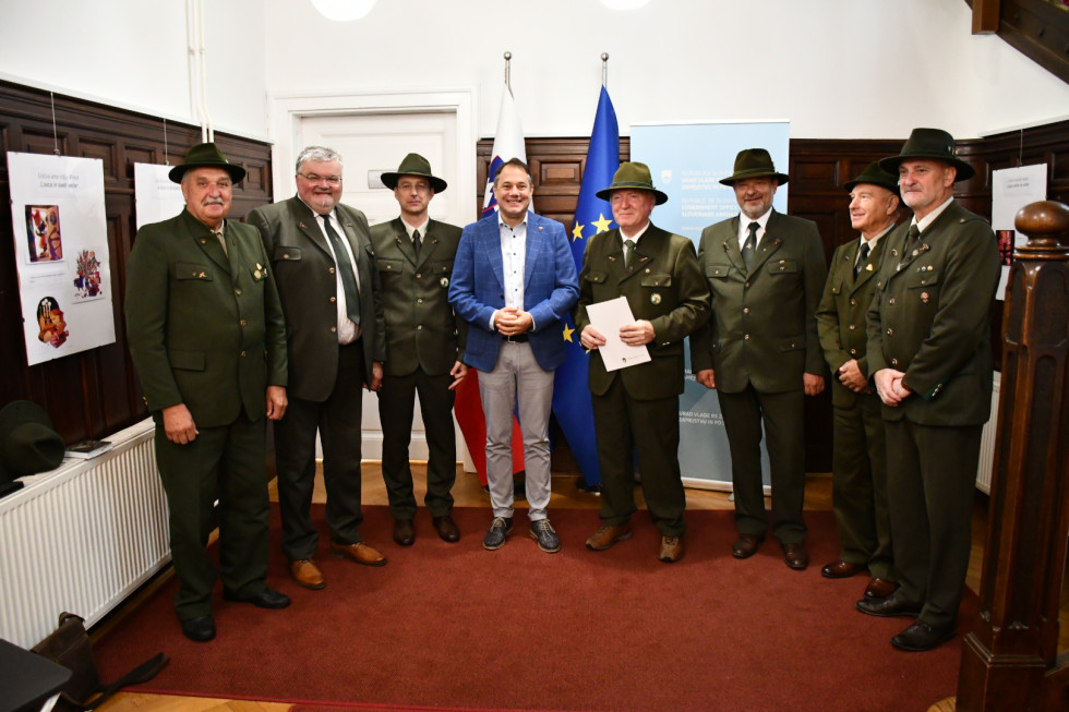 Minister in lovci pred slovensko in evropsko zastavo stojijo. Odlikovanec ima v rokah odlikovanje.